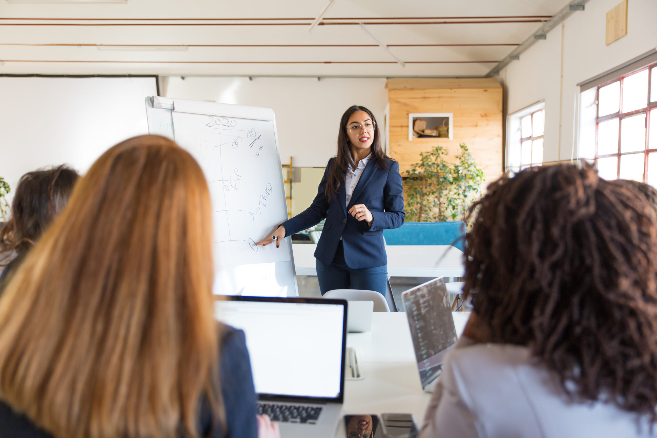 swoman pointing at whiteboard during presentation. Multiethnic female colleagues looking at speaker standing near whiteboard in a training session